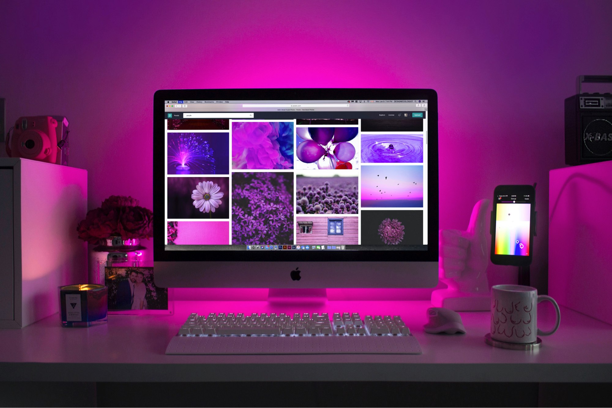 A mac computer on a desk with pink led lights with various items around it.